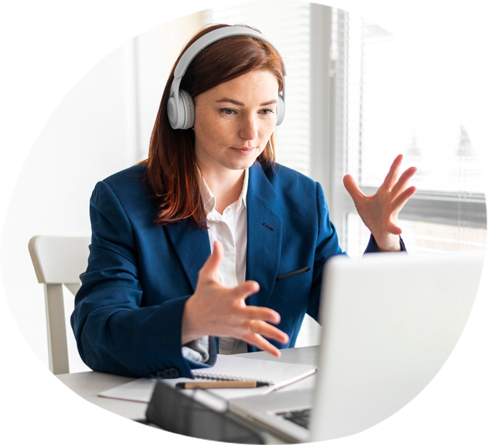 A woman working as a Virtual Assistant, wearing headphones and gesturing while engaging in a video call on her laptop. She is dressed in a blue blazer, sitting at a desk in a modern office environment.